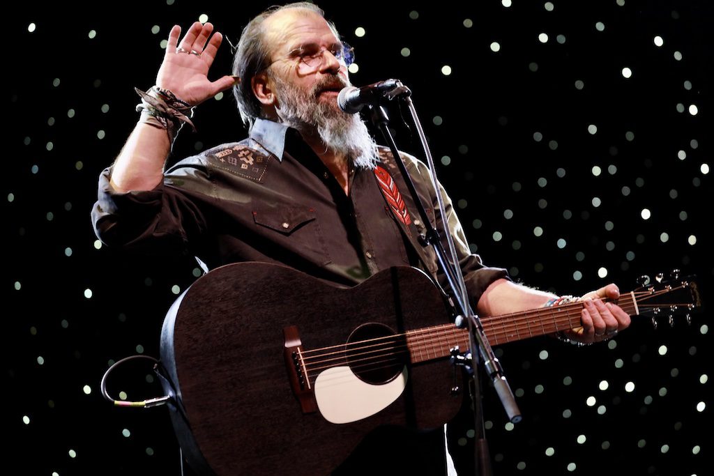 Steve Earle raises his hand as he plays an acoustic guitar