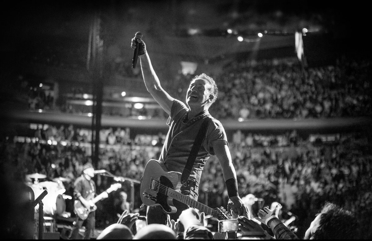 A black and white photo of Bruce Springsteen singing on stage surrounded by fans