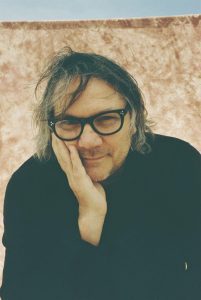A portrait of Wilco frontman Jeff Tweedy with his hand against his face, wearing glasses and a black shirt