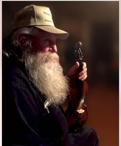 A fiddle player with white beard holds his fiddle at rest and looks to the side