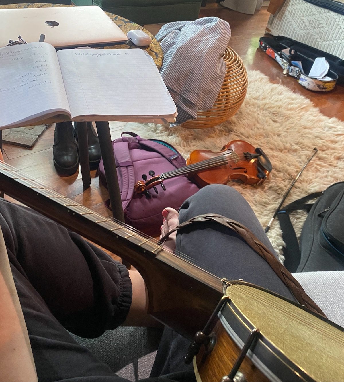 A songwriter in a living room sits with a banjo on her lap and a fiddle, notebook, and laptop computer in the background