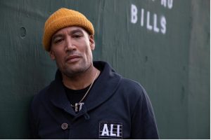 Ben Harper leans against a black wall wearing a yellow beanie hat and a black shirt that says "Ali".