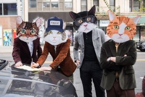 Four men wearing cat masks lean against the trunk of a car parked on the side of a city street