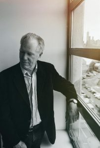 Loudon Wainwright stands by a window on a high floor