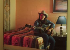 Mariel Buckley sits on a motel bed with her acoustic guitar