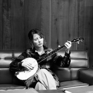 Black and white photo of Nora Brown sitting on a leather sofa playing banjo