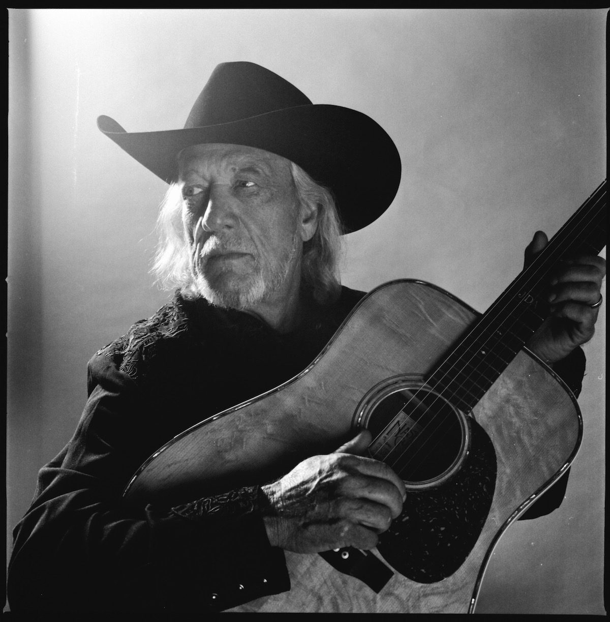 A black and white portrait of John Anderson with acoustic guitar and black hat