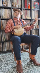 Dom Flemons playing a banjo in front of his vinyl collection