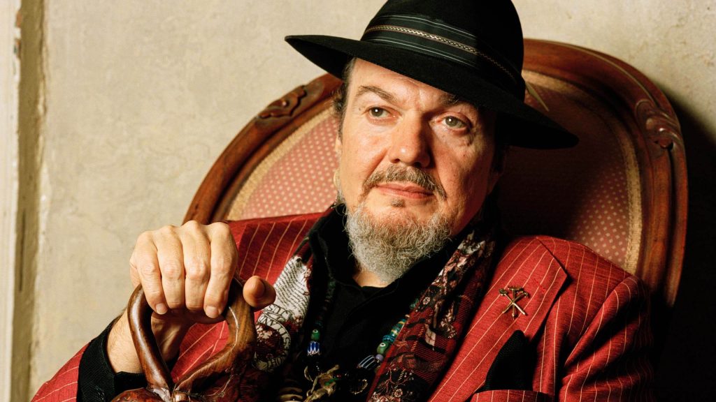 Dr. John in red jacket and black hat seated in a chair
