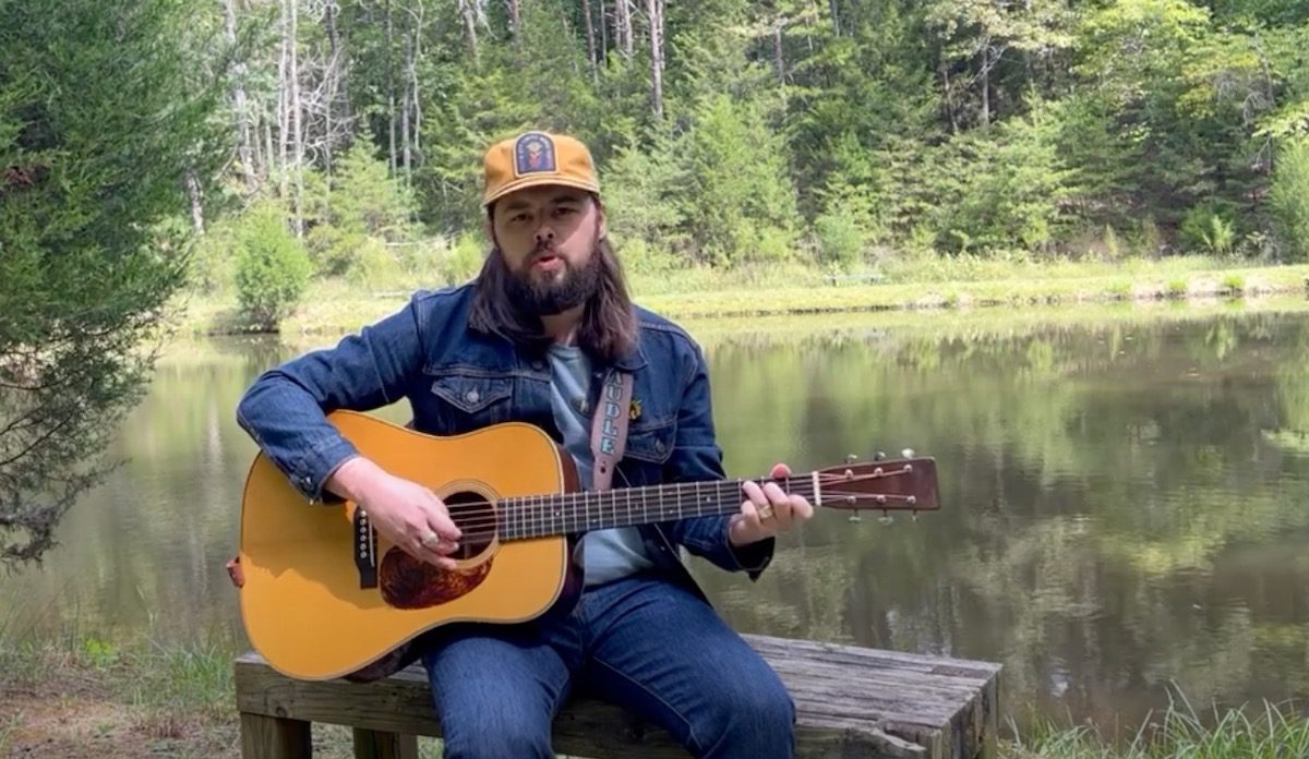 Caleb Caudle playing an acoustic guitar by a lake