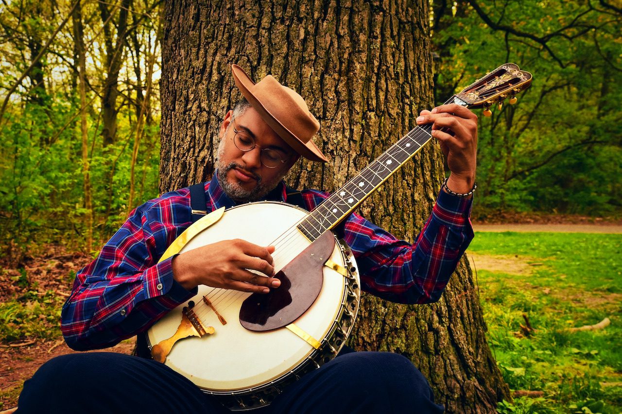 Dom Flemons plays a banjo against the trunk of a tree