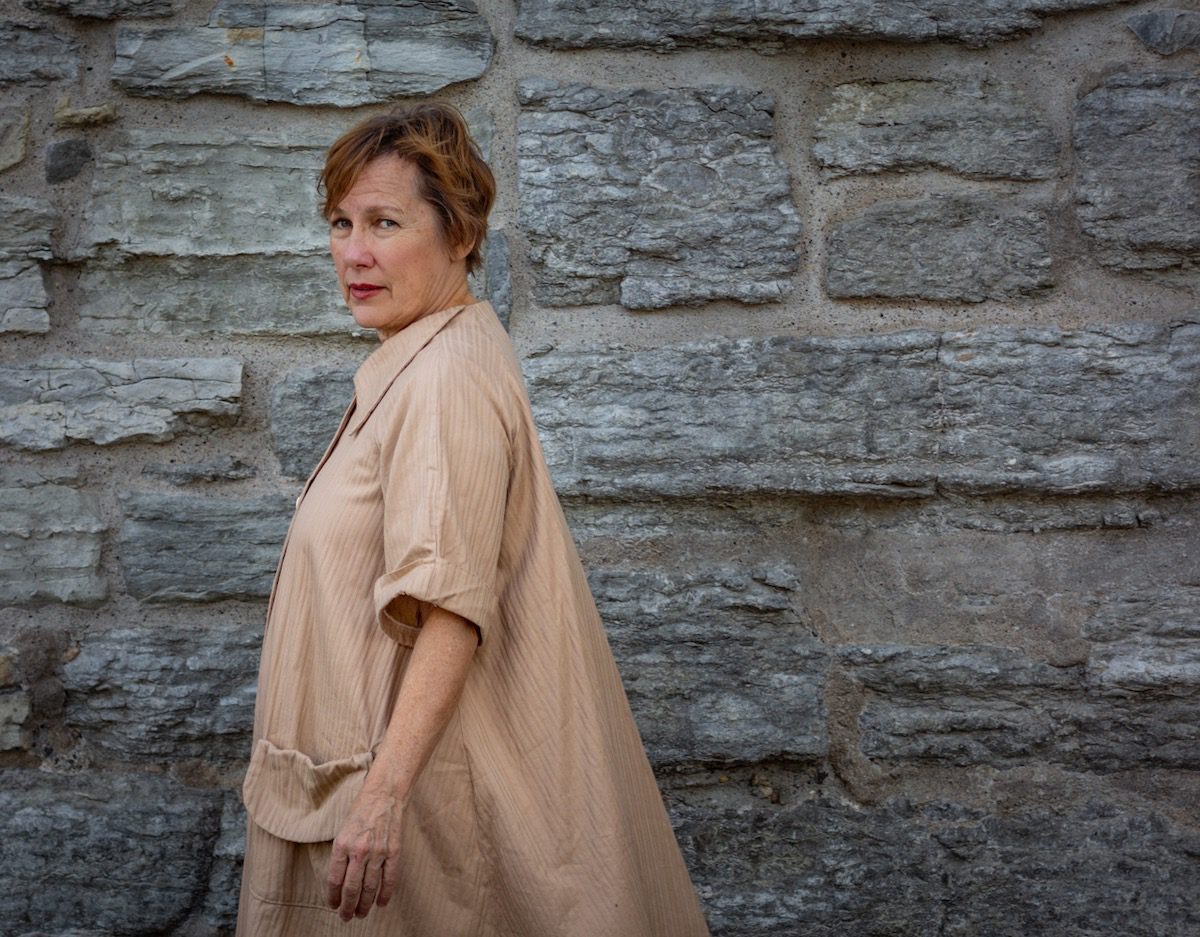 Iris DeMent side angle in a tan cloak against a stone wall