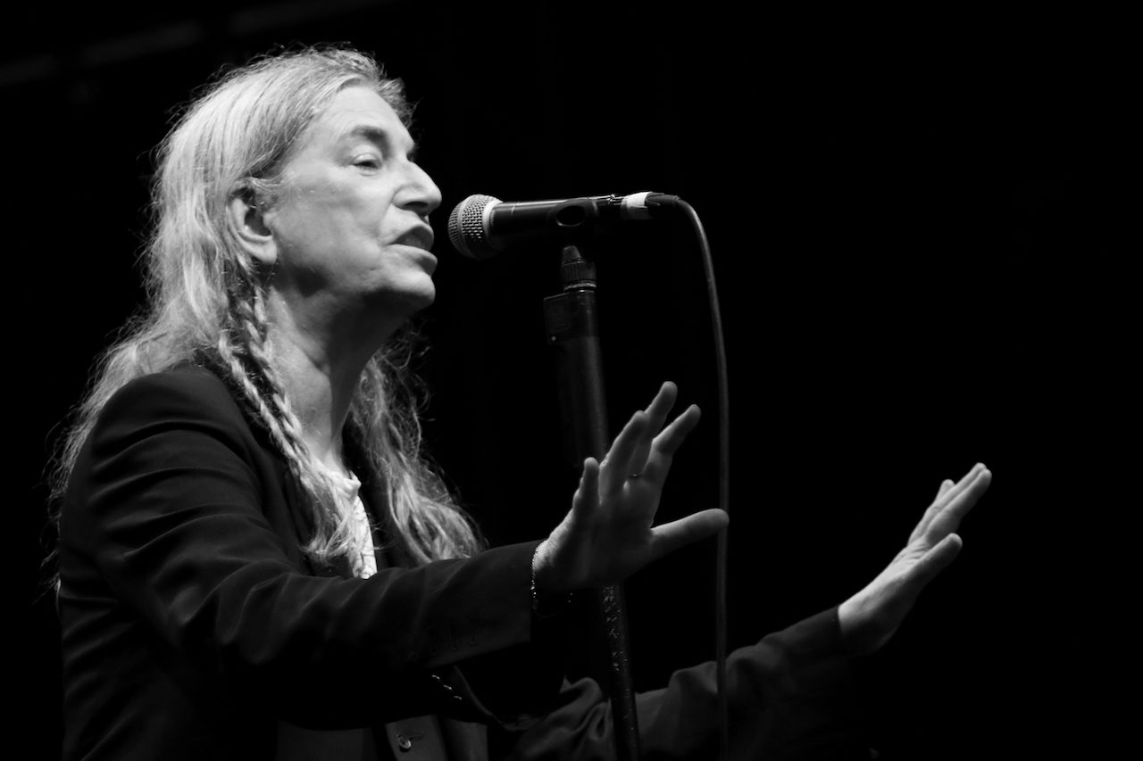 Black and white portrait of Patti Smith at the microphone, her silver hair braided
