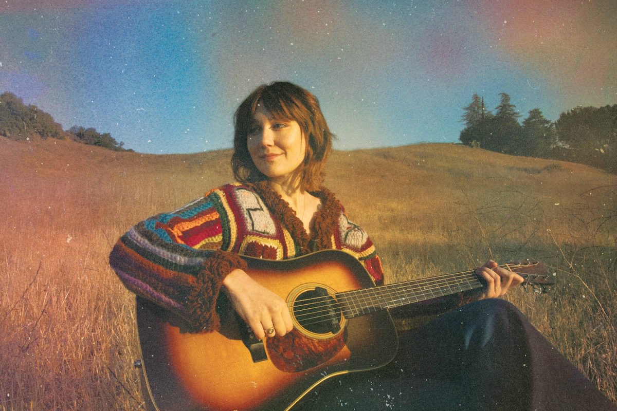 Molly Tuttle sits in a field with an acoustic guitar.