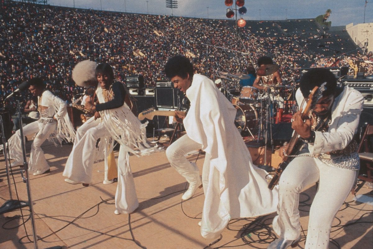 The Bar-Kays band in white jumpsuits performs onstage at a stadium on a sunny day.