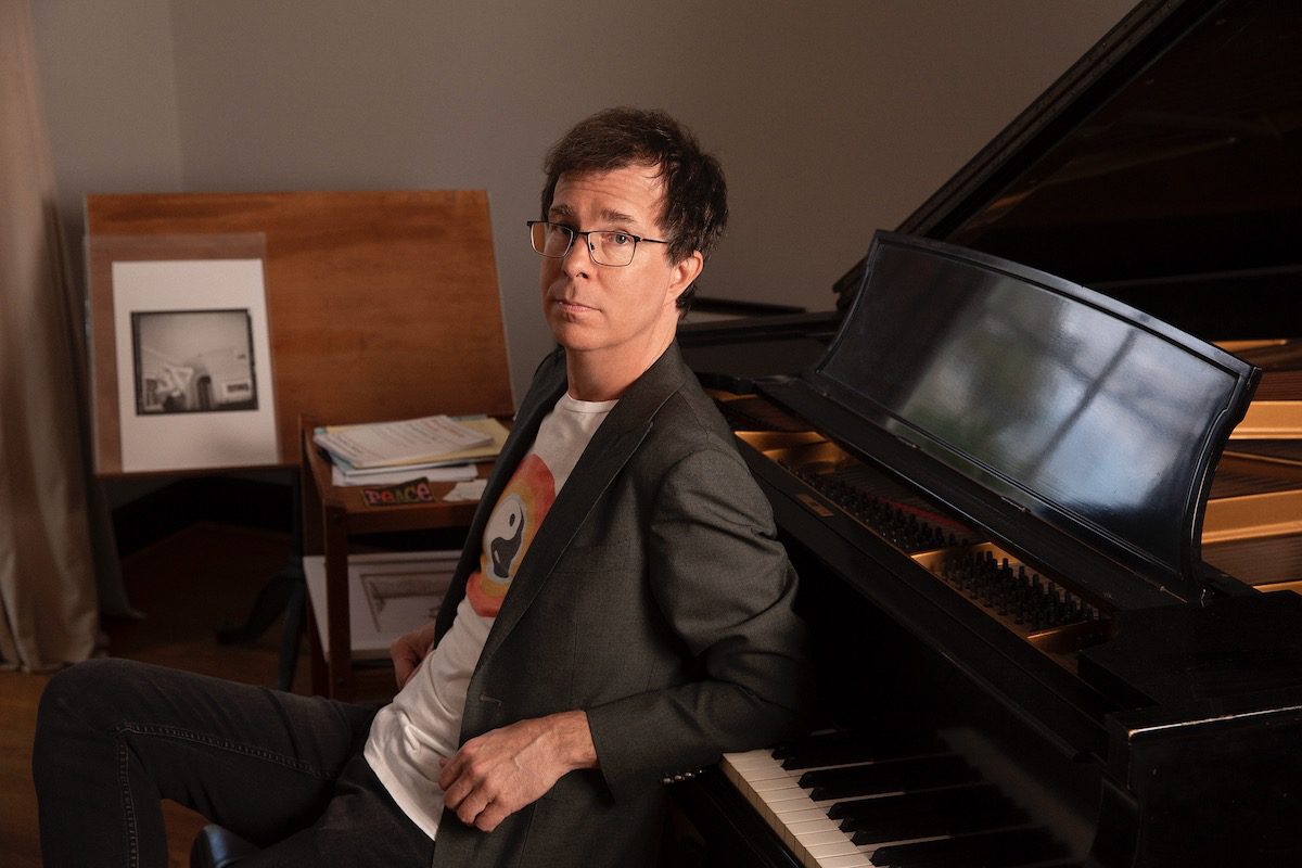 Ben Folds poses with his back to a black grand piano