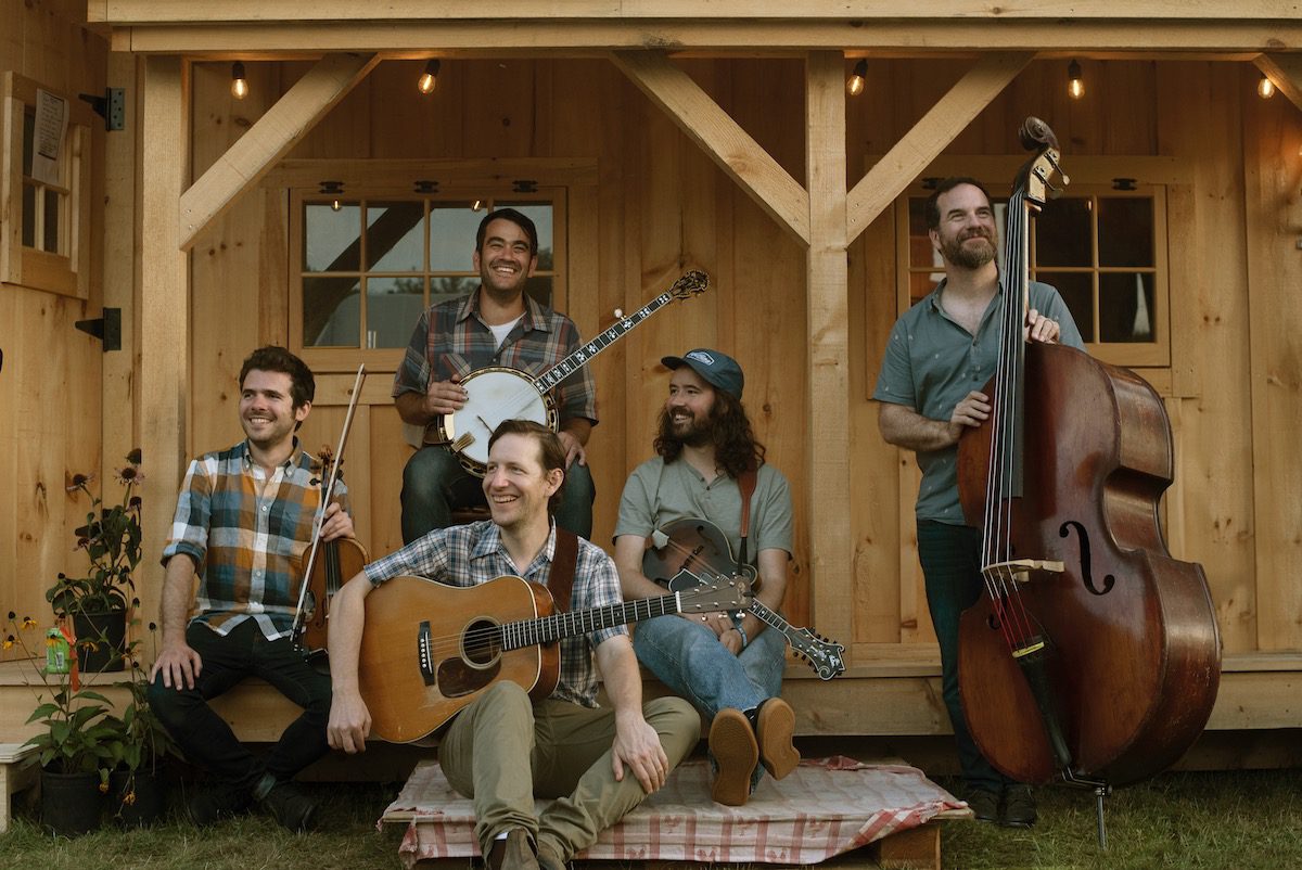 Men with bluegrass instruments sit or stand on a wooden porch