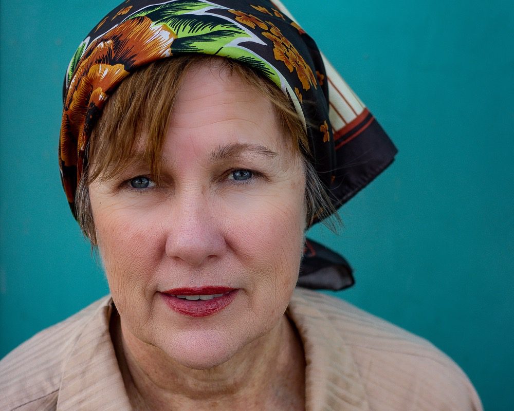 Iris DeMent against a teal background wearing a colorful scarf in her hair