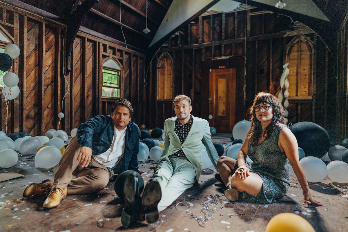 The three members of Nickel Creek sit on the wood floor of an unfinished wood building surrounded by balloons and confetti