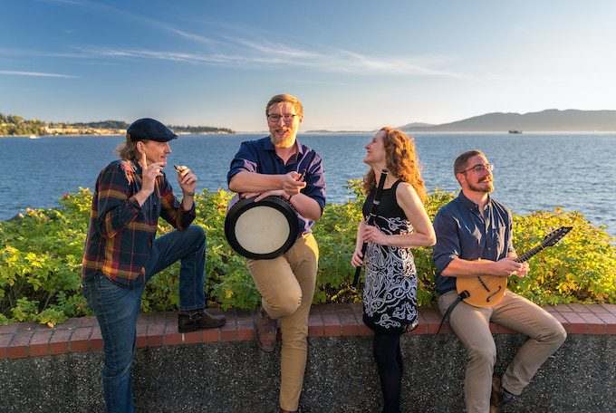 A quartet of musicians sits on a wall overlooking a golden hour seascape.