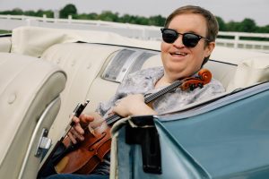 Michael Cleveland holds his fiddle in his lap in the backseat of a teal convertible with white seats.