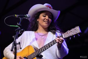 Emily Nenni in white shirt and white cowboy hat playing an acoustic guitar onstage