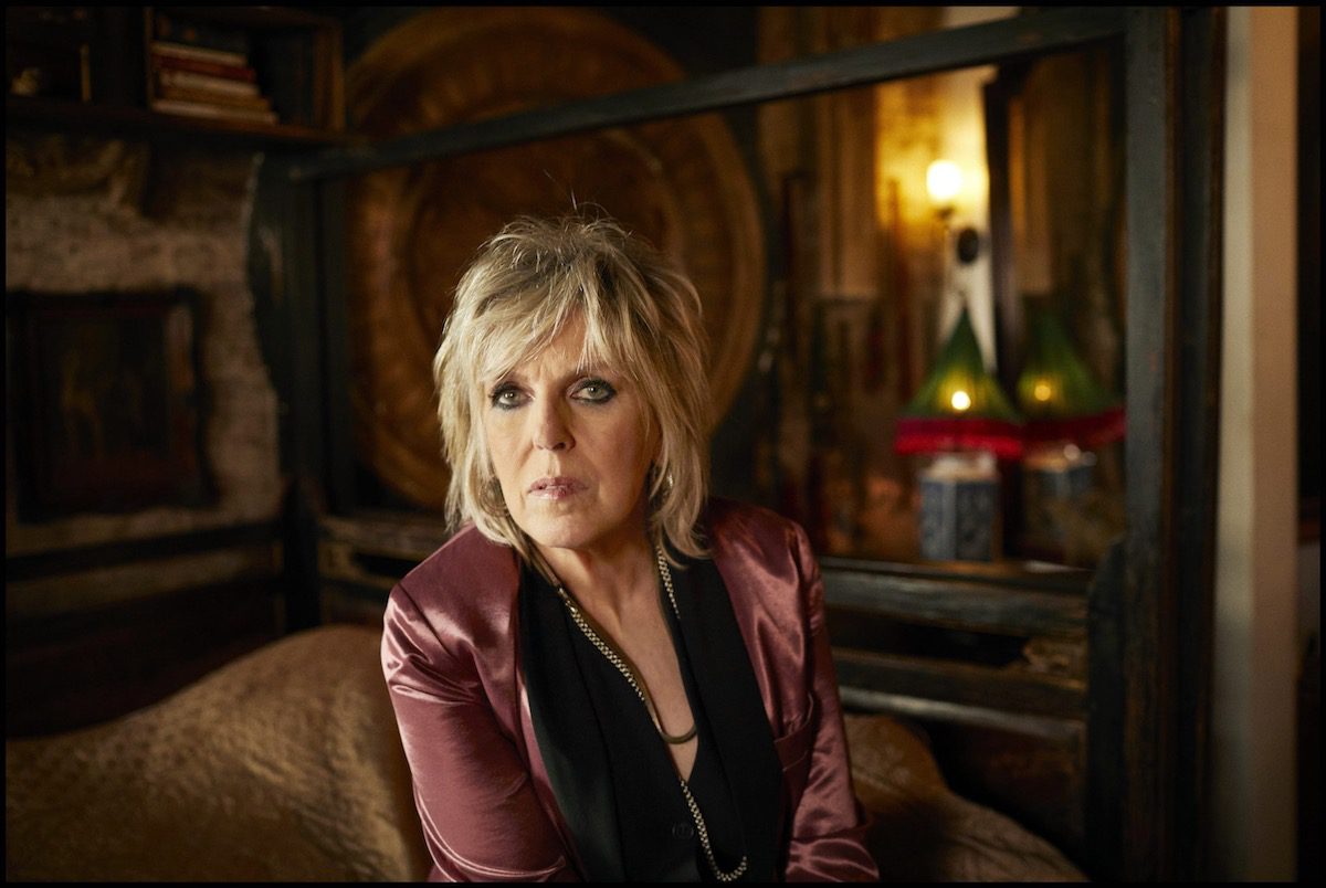 Lucinda Williams in pink jacket in a dimly lit room