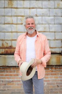 Robert Earl Keen in pink shirt and jeans stands against a brick wall holding a white cowboy hat