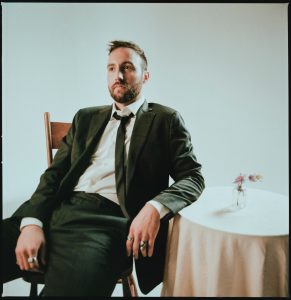 Ruston Kelly in a suit sits at a small round table covered in a white cloth