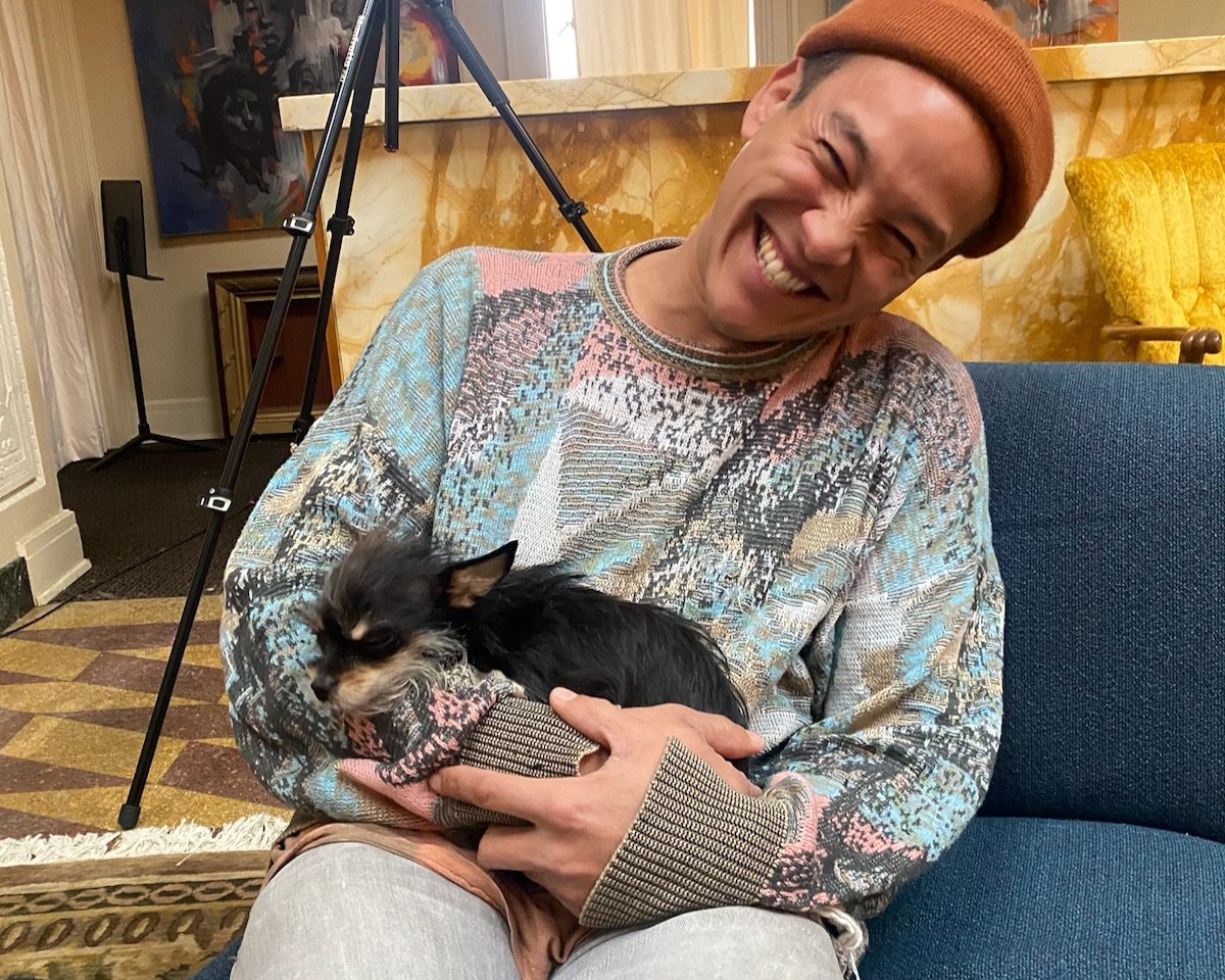 Sam Luna laughs as he sits on a sofa holding a small black and brown dog