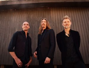 The three members of the Wood Brothers dressed all in black against a wooden wall