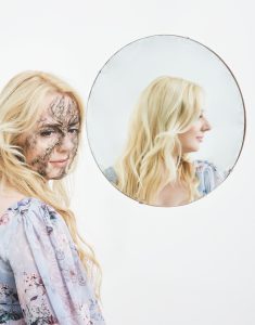 Hannah Rose Platt looks away from her image in a mirror, with black webbing creeping over her face. Her face in the mirror is clear.