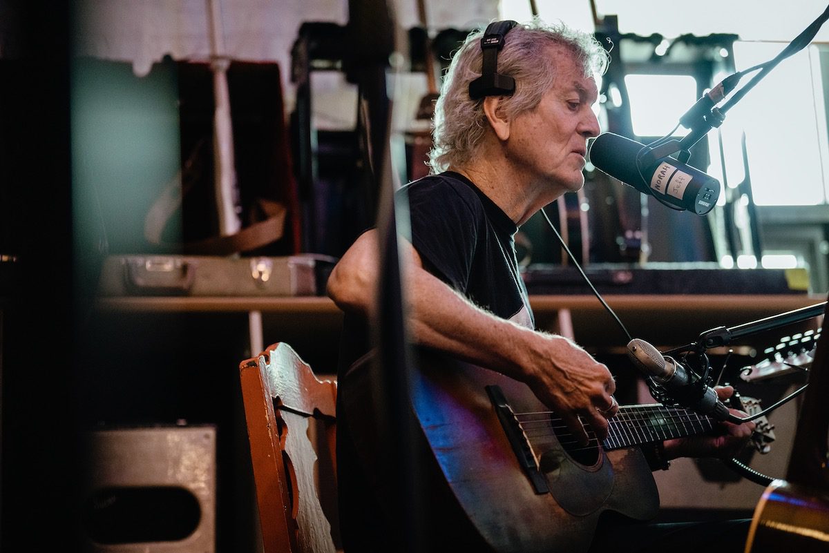 Rodney Crowell plays an acoustic guitar in a studio amid lots of gear