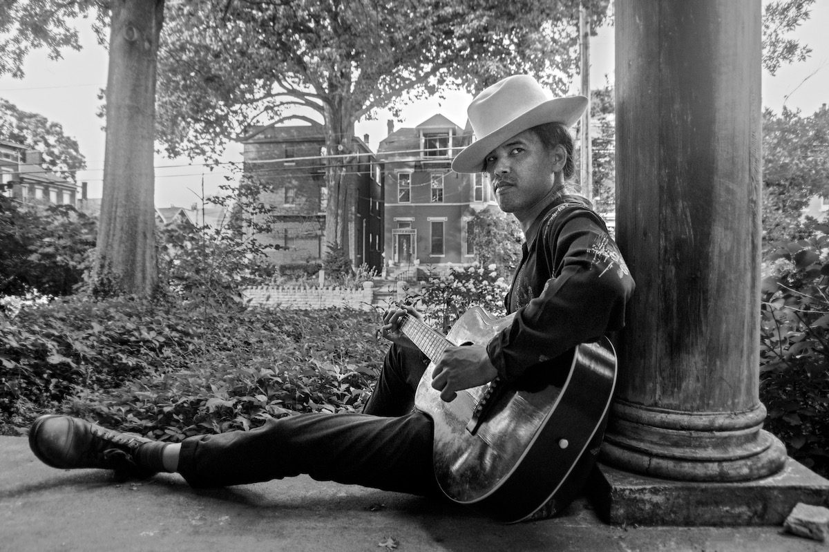 Nat Myers plays guitar while leaning against the column of a building