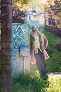 Rufus Wainwright in a fuzzy coat leaning against a painted metal box with a grassy background