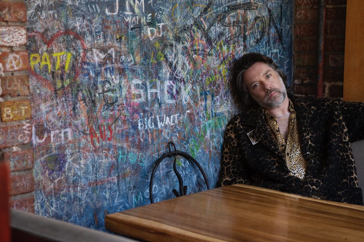 Rufus Wainwright seated at a wooden table with his head against a graffiti painted wall