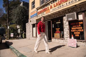 Rufus Wainwright walks past a cleaners on a city sidewalk, wearing white pants and a red shirt