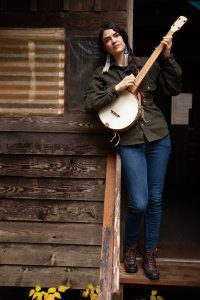 Annie Bartholomew holds a banjo as she leans against a wood framed house