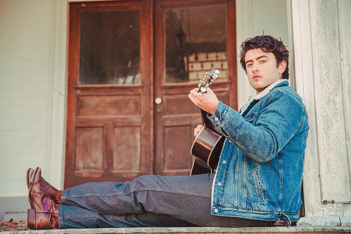 Jake Ybarra in denim jacket plays guitar on a front porch