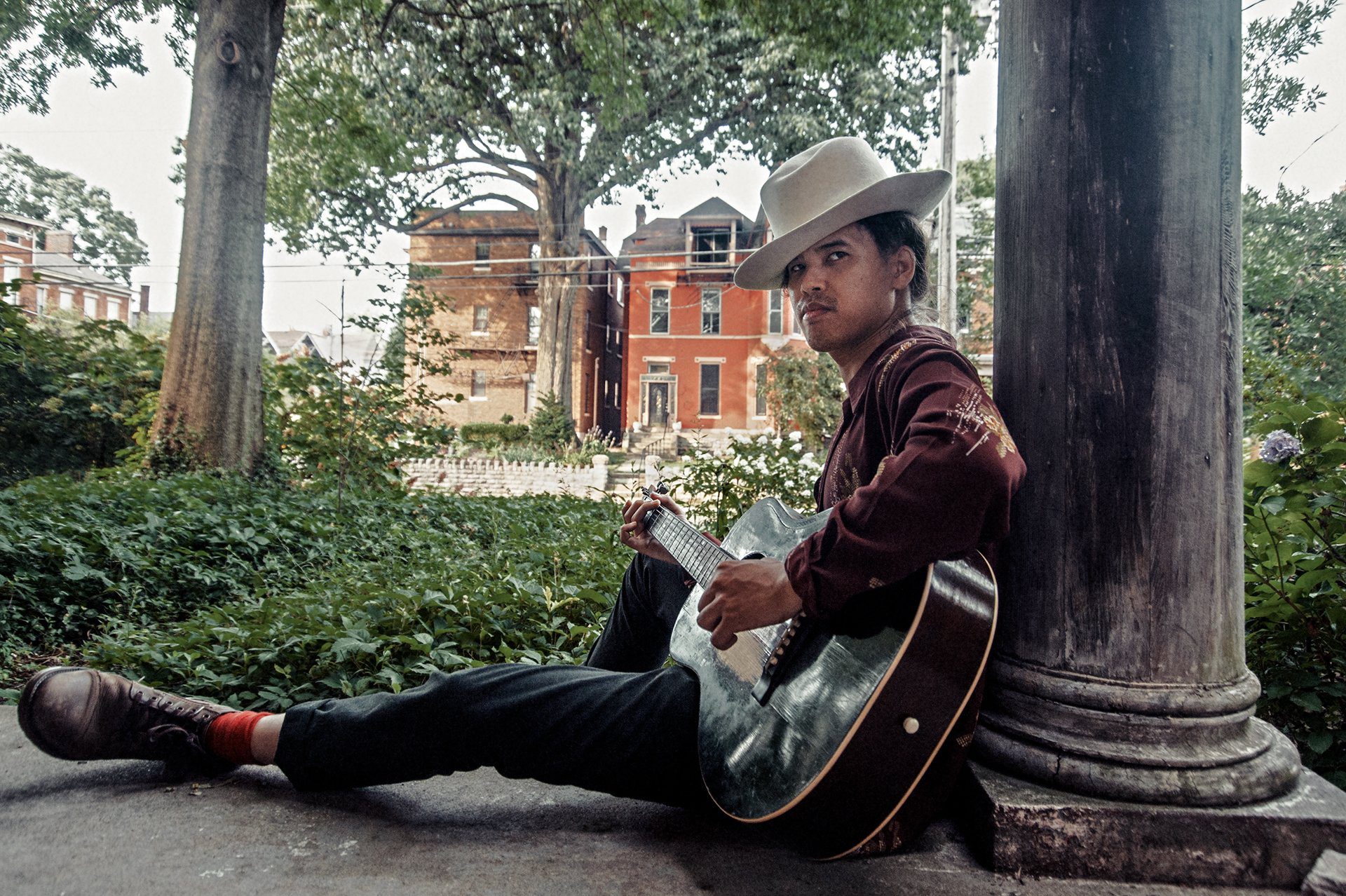Nat Myers leans against a building column and strums an acoustic guitar