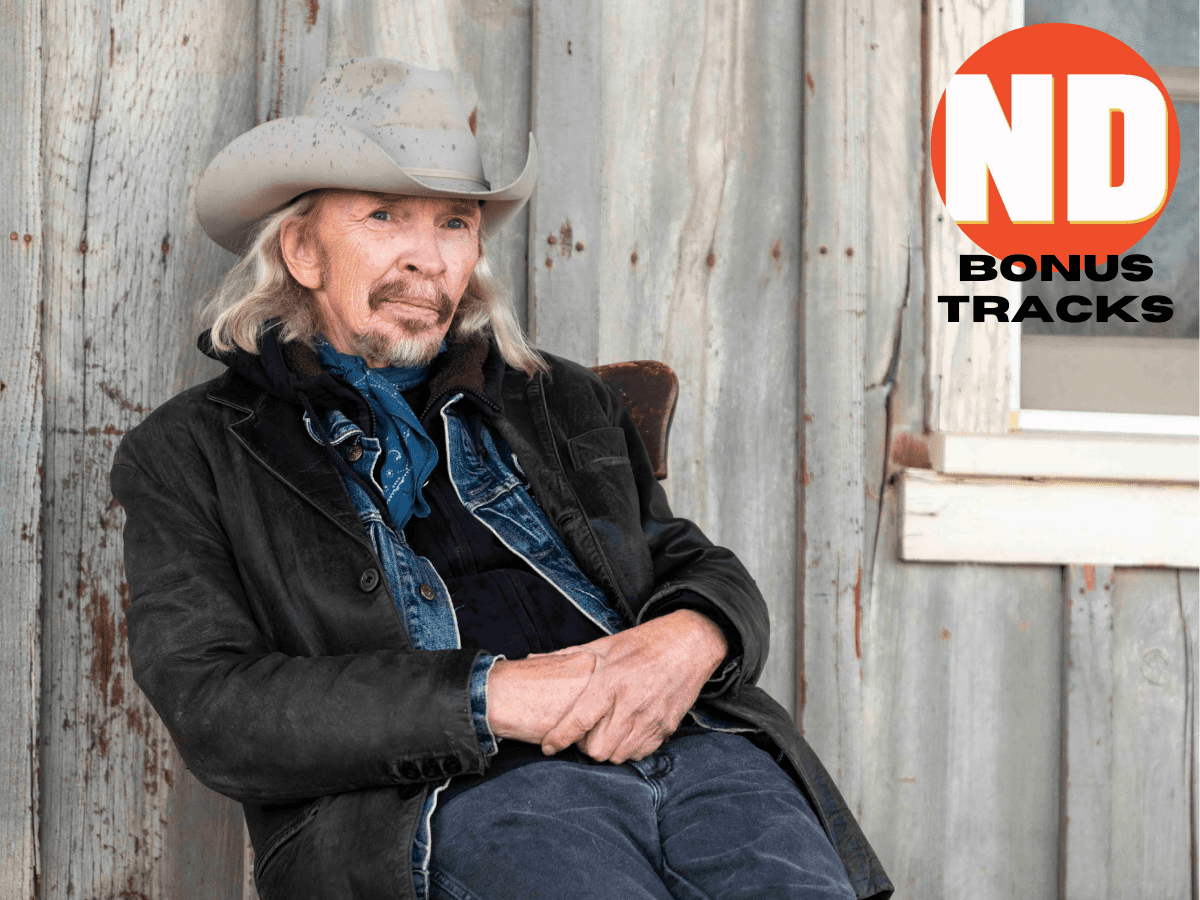 Dave Alvin seated in a chair against a grey wood wall